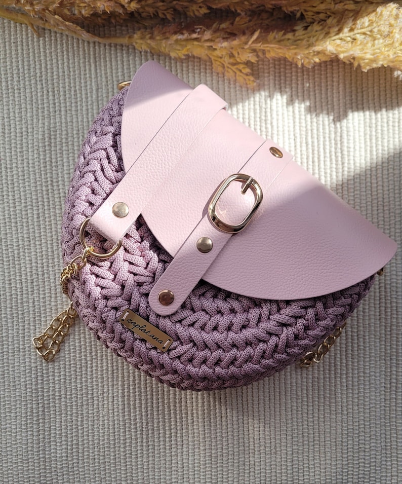 May include: A light pink crocheted handbag with a leather flap and a gold buckle. The bag has a gold chain strap and a small gold tag with the text "zaplat.ana" on it.