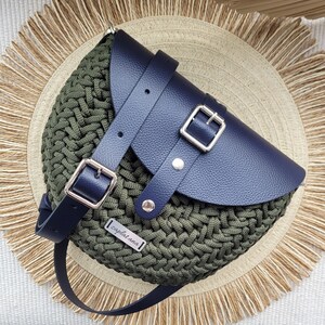 May include: A dark green crocheted bag with a navy blue leather flap and a silver buckle. The bag has a black leather strap and a silver metal tag that says "zaplatana".
