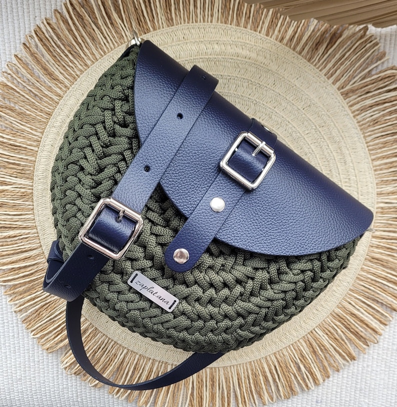 May include: A dark green crocheted bag with a navy blue leather flap and a silver buckle. The bag has a black leather strap and a silver metal tag that says "zaplatana".