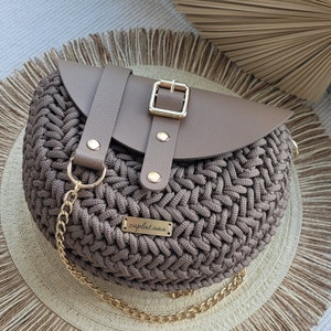 May include: A circular, woven handbag in brown tones, with a leather flap fastened by a gold buckle. It has a gold chain strap and a small metal tag that reads "zaplatana."