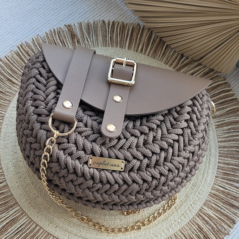 May include: A circular, woven handbag in brown tones, with a leather flap fastened by a gold buckle. It has a gold chain strap and a small metal tag that reads "zaplatana."