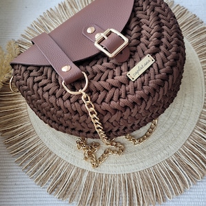 May include: A brown crocheted half-moon shaped handbag with a brown leather flap and a gold buckle. The bag has a gold chain strap and a gold metal tag that says "zaplat.ana".