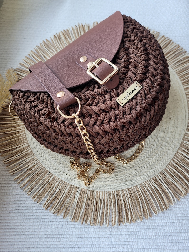 May include: A brown crocheted half-moon shaped handbag with a brown leather flap and a gold buckle. The bag has a gold chain strap and a gold metal tag that says "zaplat.ana".
