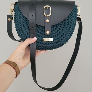 May include: A teal and black crossbody bag with a woven texture. The bag features a black leather flap secured by a gold-coloured buckle and strap. The bag has a long black strap and a small gold-coloured tag that reads "zaplatana".