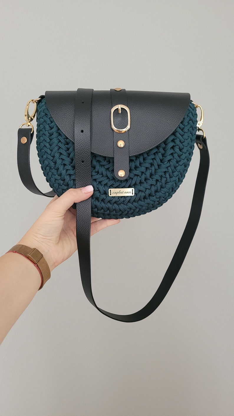 May include: A teal and black crossbody bag with a woven texture. The bag features a black leather flap secured by a gold-coloured buckle and strap. The bag has a long black strap and a small gold-coloured tag that reads "zaplatana".