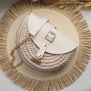 May include: A circular, beige crochet handbag with a cream leather flap, fastened with a gold buckle. It has a gold chain strap and sits on a woven mat with a fringed edge. The design is neutral and appears handmade.