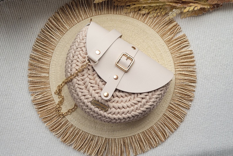 May include: A circular, beige crochet handbag with a cream leather flap, fastened with a gold buckle. It has a gold chain strap and sits on a woven mat with a fringed edge. The design is neutral and appears handmade.