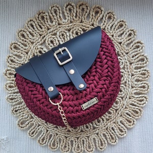 May include: A burgundy crocheted handbag with a black leather flap and gold buckle. The bag has a gold chain strap and a small metal tag reading "zaplat ana". It rests on a woven circular placemat.