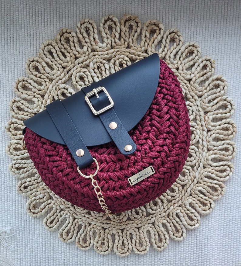 May include: A burgundy crocheted handbag with a black leather flap and gold buckle. The bag has a gold chain strap and a small metal tag reading "zaplat ana". It rests on a woven circular placemat.