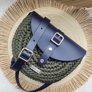 May include: Olive green crocheted crossbody bag with a navy blue leather flap and strap. The bag features silver buckles and a small rectangular tag with the word "zaplatana". The bag is set on a woven straw mat.
