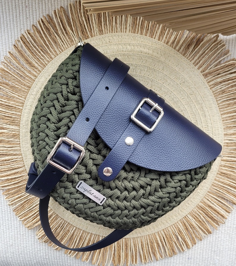 May include: Olive green crocheted crossbody bag with a navy blue leather flap and strap. The bag features silver buckles and a small rectangular tag with the word "zaplatana". The bag is set on a woven straw mat.