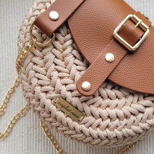May include: A beige crocheted handbag with a brown leather flap and strap. The bag has a gold chain strap, gold hardware, and a small gold tag that reads "zapl.at.ana."