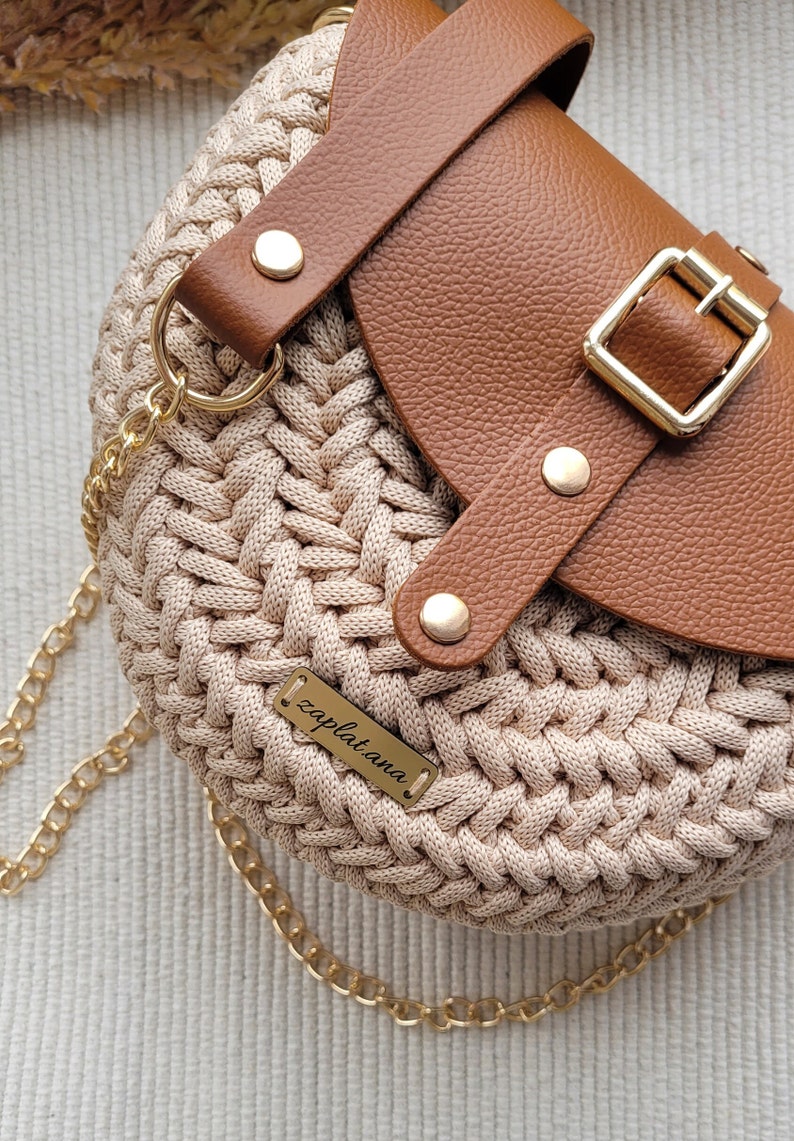 May include: A beige crocheted handbag with a brown leather flap and strap. The bag has a gold chain strap, gold hardware, and a small gold tag that reads "zapl.at.ana."