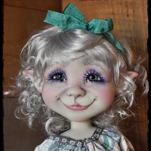 May include: A doll with silver hair and a teal bow, wearing a white dress with a black and teal pattern. The doll has large blue eyes and a painted smile.