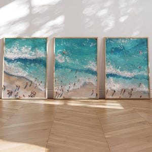 May include: Three vertical framed wall art prints featuring an aerial view of a sandy beach and turquoise ocean waves. The textured paintings depict small figures of people swimming in the water and walking along the shoreline in bright light.
