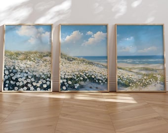 May include: A set of three framed wall art prints featuring a continuous coastal landscape. Each print displays white flowers blooming on sandy dunes under a bright blue sky with soft white clouds, overlooking a calm ocean horizon.