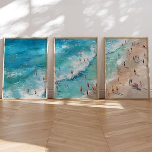 Pode incluir: Um conjunto de três impressões de arte de parede verticais emolduradas que retratam vistas aéreas de uma cena de praia. As pinturas apresentam tons de água do mar azul e turquesa com ondas brancas e numerosas pequenas figuras de pessoas.