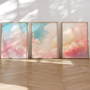 May include: Three rectangular wall art prints in light wooden frames, each displaying a soft, painterly depiction of clouds in a sky with gradients of pastel pink, peach, and light blue, arranged side by side on a light-colored floor.