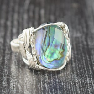 May include: A handcrafted ring featuring a large, iridescent abalone shell gemstone. The gemstone is a mix of blues, greens, and purples, set in a woven silver-toned metal band. The ring is displayed on a dark wooden surface.