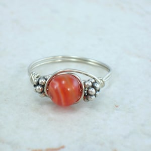 May include: A silver wire ring with a red and white agate bead. The bead is surrounded by small silver beads.