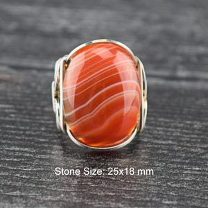 May include: A ring featuring a large, oval-shaped, orange and white banded agate stone set in a silver-toned wire-wrapped metal band. The stone has a smooth, polished surface with visible natural layers. The text Stone Size: 25x18 mm is present.