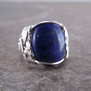 May include: A ring featuring a large, oval-shaped, deep blue lapis lazuli gemstone with subtle gold-colored flecks. The stone is secured by a decorative, intricate silver-colored wire-wrapped setting that forms the band and surrounds the gem.
