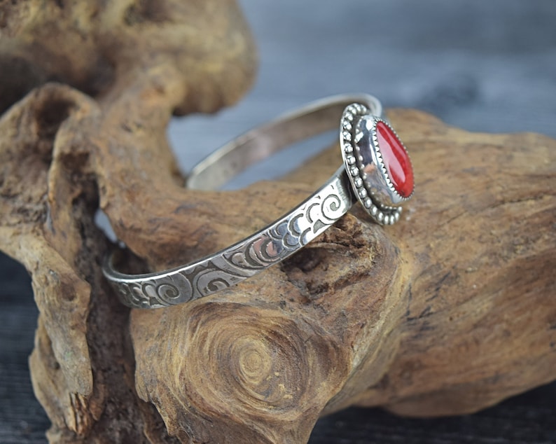 May include: A silver bangle bracelet with an ornate design and a red gemstone set in a silver bezel.