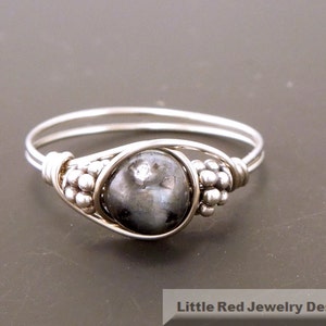 May include: A silver ring with a black gemstone set in a wire-wrapped design. The ring has a beaded detail on the band.