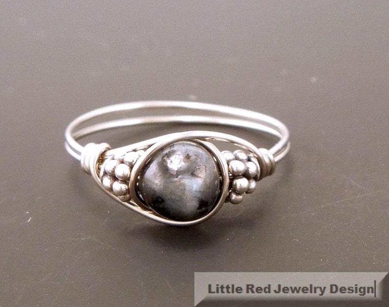 May include: A silver ring with a black gemstone set in a wire-wrapped design. The ring has a beaded detail on the band.