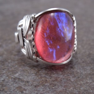 May include: A silver-toned ring featuring a large, oval-shaped, iridescent gemstone. The gemstone displays a blend of red and blue hues. The ring's band is intricately woven with silver wire, adding a unique texture to the jewelry.