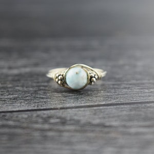 May include: A silver-toned ring with a light blue, round gemstone at the center. The ring features a wire-wrapped design with small, decorative bead clusters on either side of the gemstone. The ring is set against a dark, wooden background.