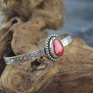 May include: A silver bracelet with a red coral stone set in a silver bezel. The bracelet has a swirling pattern.