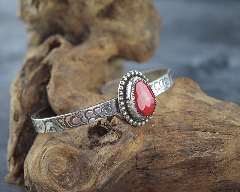 May include: A silver bracelet with a red coral stone set in a silver bezel. The bracelet has a swirling pattern.