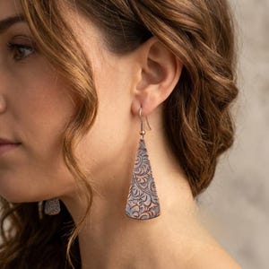 May include: A pair of dangling earrings featuring a triangular, copper-toned metal plate with a dark, recessed floral pattern. The earrings hang from a simple metal hook wire, showcasing a textured surface with a mix of metallic copper and dark gray tones.