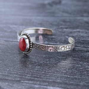 May include: A silver bracelet with a red gemstone set in a bezel setting. The bracelet has a delicate, swirling design.