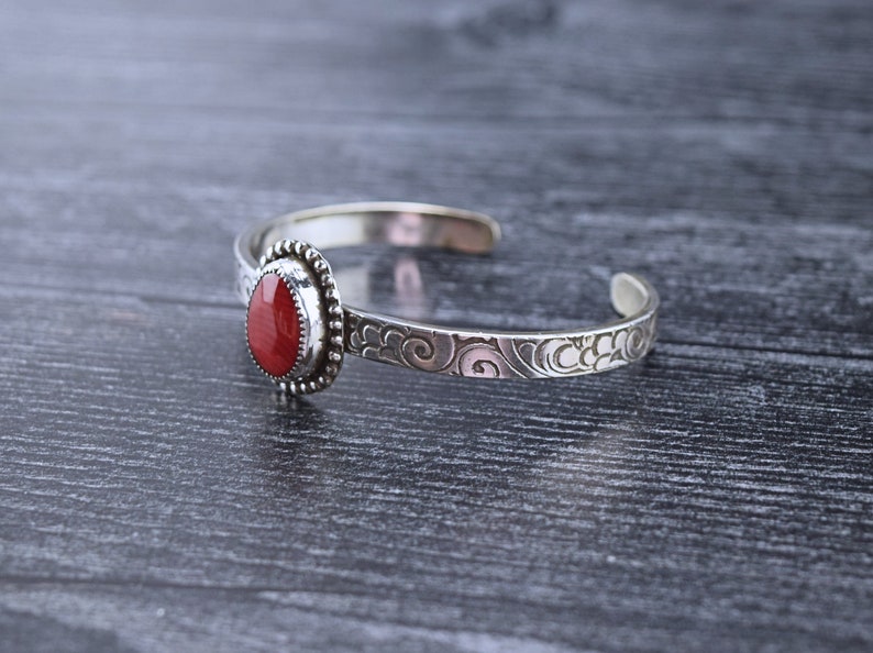 May include: A silver bracelet with a red gemstone set in a bezel setting. The bracelet has a delicate, swirling design.