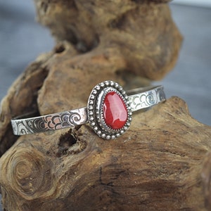 May include: A silver bangle bracelet with an ornate design and a red gemstone set in a silver bezel.