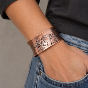 May include: A wide copper cuff bracelet featuring a detailed embossed design of multiple horses running in a line. The metal has a warm, reddish-brown metallic finish with darkened recessed areas that highlight the intricate equine silhouettes.