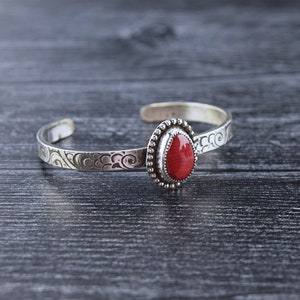 May include: A silver bracelet with a red stone set in a silver bezel. The bracelet has a decorative design on the band.