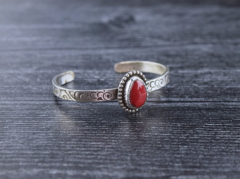 May include: A silver bracelet with a red stone set in a silver bezel. The bracelet has a decorative design on the band.