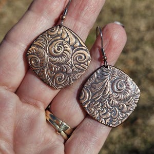 May include: Copper-colored, diamond-shaped earrings with intricate embossed floral patterns. The earrings have a vintage aesthetic, featuring a detailed design of swirling leaves and curls. The ear wires are silver-toned with a small copper bead.