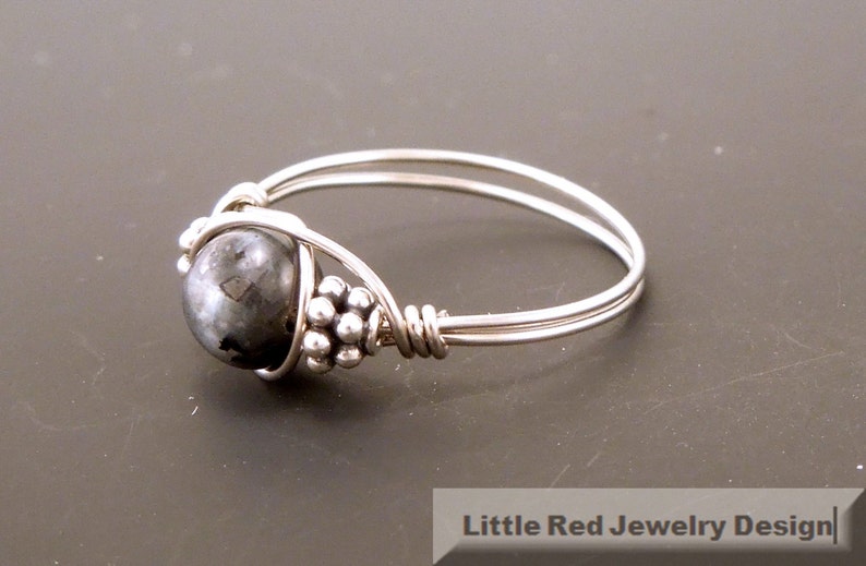 May include: A silver ring with a black gemstone wrapped in wire. The ring is set with small silver beads.