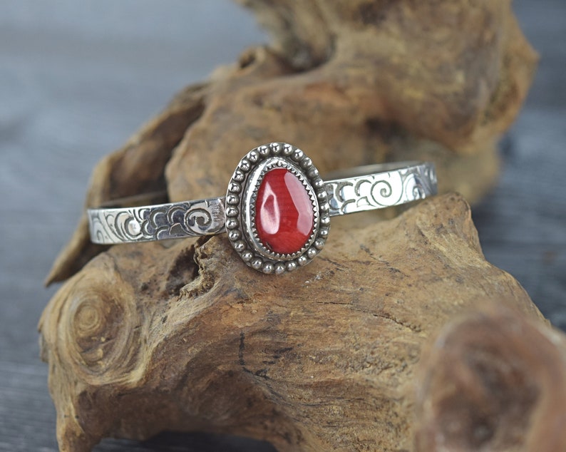 May include: A silver bangle bracelet with an ornate design and a red gemstone set in a silver bezel.