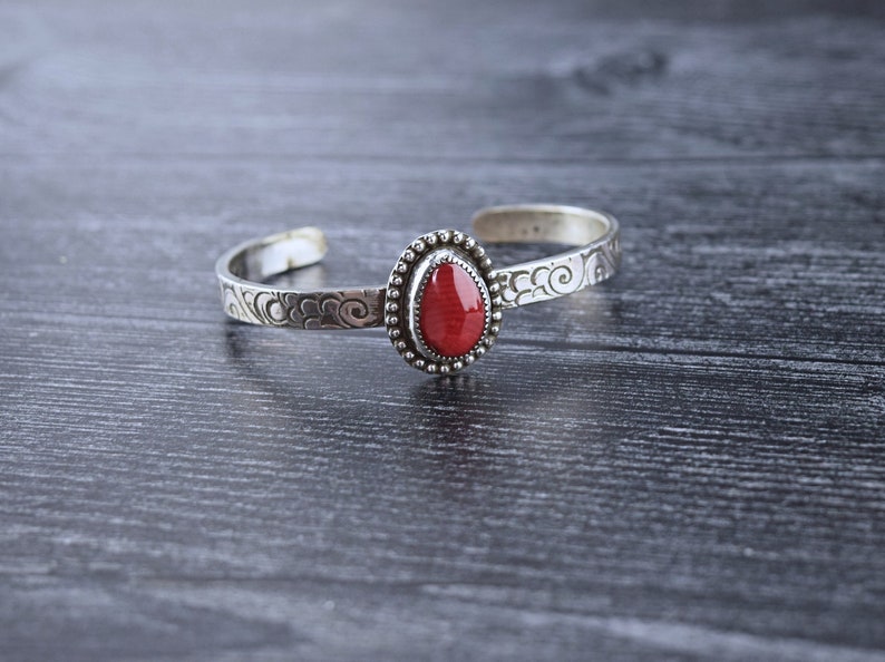 May include: A silver bracelet with an ornate design and a red gemstone. The bracelet is on a dark gray wooden surface.