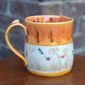 May include: A ceramic mug with a light orange glaze and a white band featuring a dragonfly and butterfly design. The mug has a rounded body and a handle.