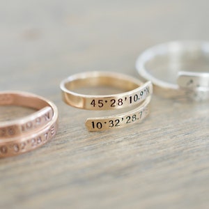 May include: Three gold, silver, and rose gold rings with coordinates engraved on them. The rings are stacked on a wooden surface.