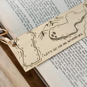 May include: A beige bookmark featuring a hand-drawn map design and the words "LET'S GO ON AN ADVENTURE." The bookmark rests on an open book, with gold-coloured hardware.