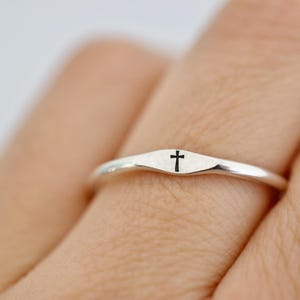 Könnte beinhalten: Ein silberner Ring mit einem kleinen, eingravierten schwarzen Kreuz. Der Ring hat ein schlichtes, elegantes Design mit glatter, polierter Oberfläche. Das Kreuz ist mittig auf der Ringoberfläche platziert und macht es zu einem dezenten, aber aussagekräftigen Schmuckstück.