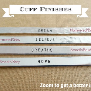 May include: Four silver metal cuff bracelets with different finishes. The top bracelet has the word "DREAM" engraved on it and is hammered with a brushed finish. The second bracelet has the word "BELIEVE" engraved on it and is hammered with a shiny finish. The third bracelet has the word "BREATHE" engraved on it and is smooth with a brushed finish. The bottom bracelet has the word "HOPE" engraved on it and is smooth with a shiny finish.