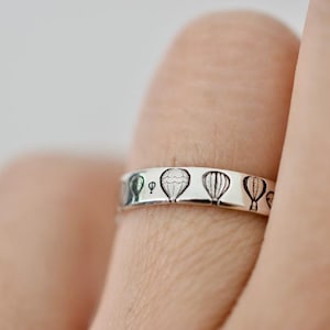 Puede incluir: Un anillo de plata con un diseño de globos aerostáticos grabados alrededor de la banda.